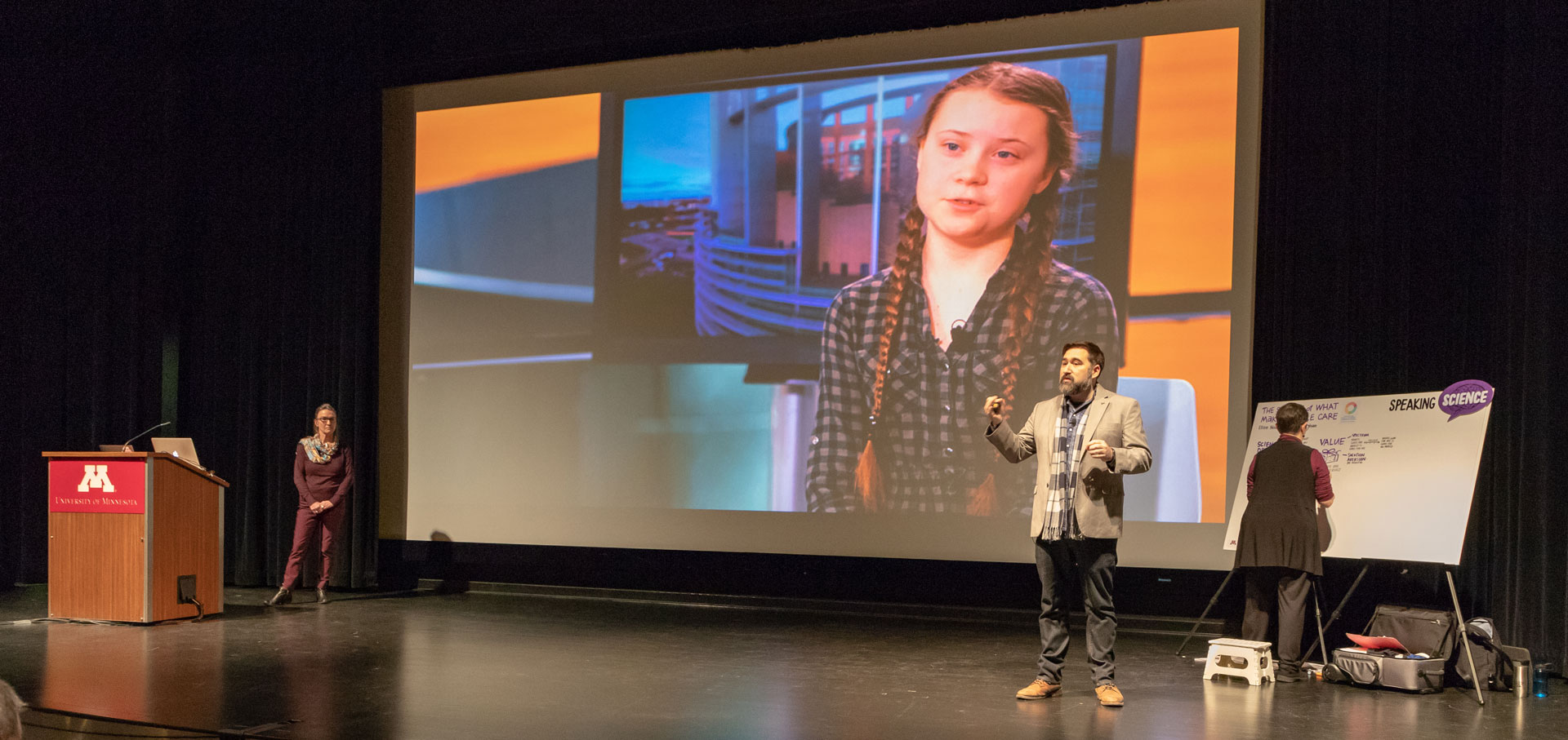  A speaker stands on stage at Speaking Science 2020. A visual of environmental activist Greta Thunberg appears on the screen behind them.