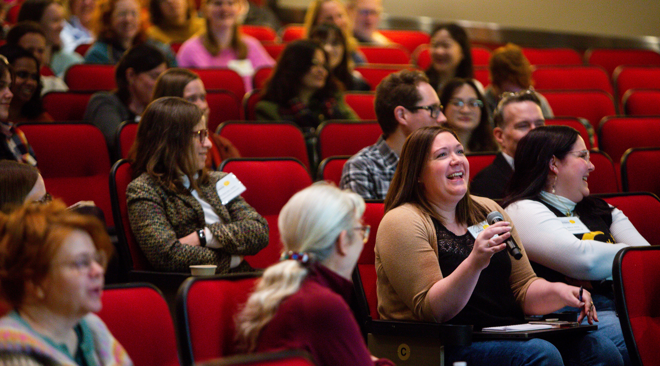 A crowd of Speaking Science attendees seated in an auditorium. One attendee holds a microphone as part of a Q & A.
