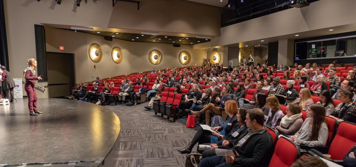 Speaking Science 2020: A person on a stage stands in front of a room full of people gathered in a large theater